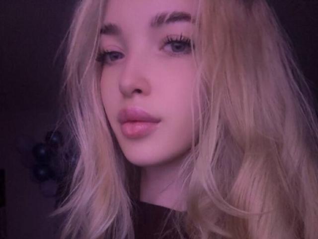 Brooke Zapf, blonde avec des cheveux ondulés, un éclairage rosâtre, un maquillage subtil et des lèvres roses, regarde légèrement sur le côté avec une expression douce et confiante. Ses cils sont longs et foncés..