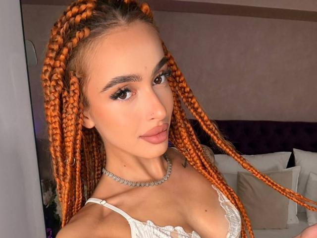 MarissaFex with orange braids, pouty lips, and a necklace, wearing a white lace top, showing her tits and a slim neck. Brown eyes, eyebrows, and subtle makeup.