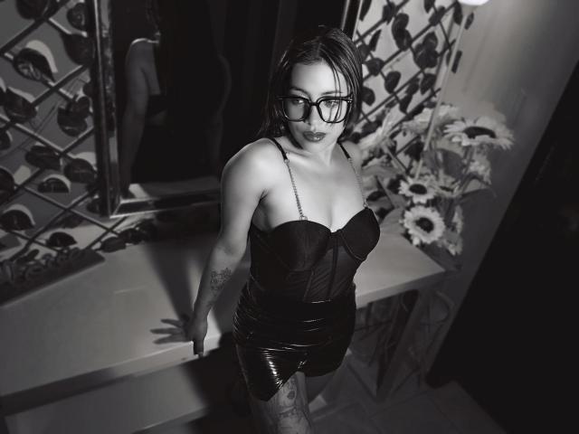 Grace Cooper mit Brille, trägt ein schwarzes, trägerloses Korsett und glänzende schwarze Shorts, steht in einem dunklen Raum, die Arme auf einem Tisch, blickt nach oben, Tattoos sind auf ihrem Arm sichtbar..