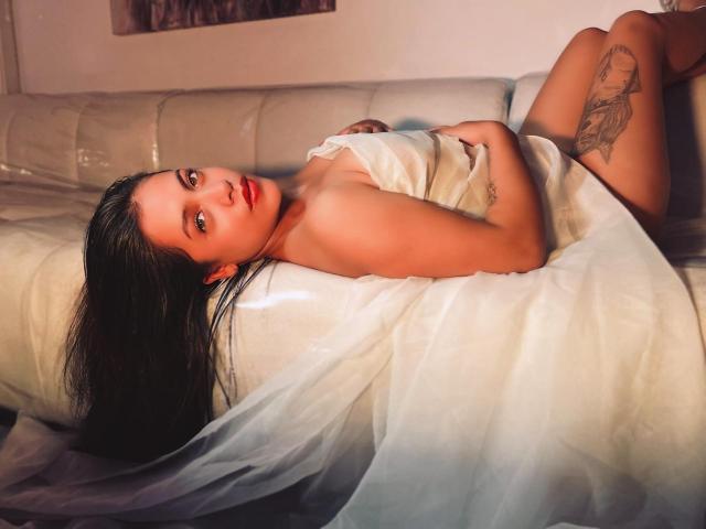 Grace Cooper deitada em um sofá branco, nua, com um lençol branco cobrindo seus seios, olhando para a câmera com batom vermelho, cabelo preto e uma tatuagem na coxa direita..