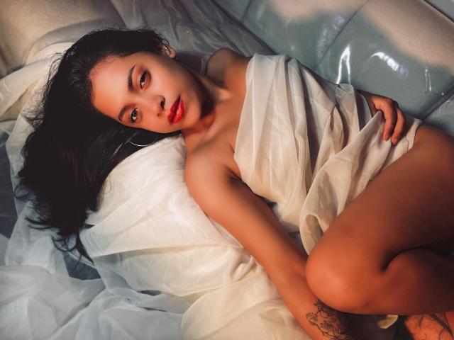 Grace Cooper liegt auf weißen Laken, in ein durchsichtiges Tuch gehüllt, wobei ihre Brüste und ihr Hintern sichtbar sind, mit rotem Lippenstift, dunklem Haar und einem Tattoo an ihrem rechten Oberschenkel..