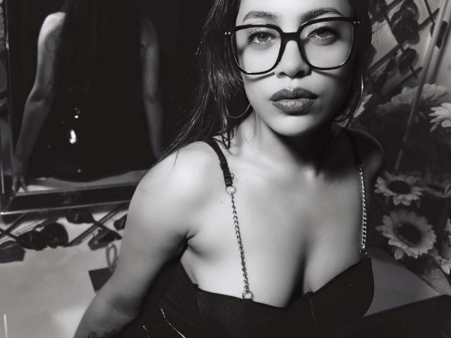 Grace Cooper, con gli occhiali, il rossetto scuro e gli orecchini a cerchio, mostra il seno in una maglietta nera, riflessa in uno specchio, con carta da parati floreale e girasoli sullo sfondo..
