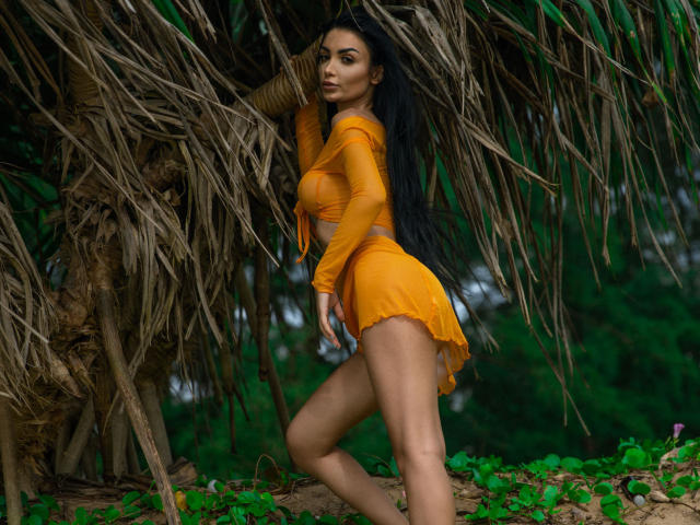 RaqueleDiva mit langen schwarzen Haaren, einem orangefarbenen Crop-Top und Shorts, steht vor einer Palmen, wobei sie ihre Brüste und ihren Hintern zur Schau stellt, im Hintergrund sind grüne Blätter und Pflanzen zu sehen..