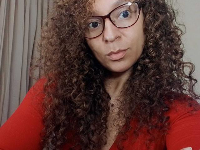 Angelonix, met krullend bruin haar, rode bril en een rood shirt, trekt lichtjes haar mondhoeken naar beneden en laat op een selfie haar borsten zien. Haar blauwe ogen vallen op tegen haar lichtbruine huid..