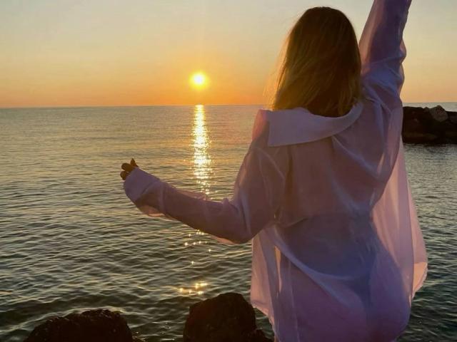 MissiSofiya with her back to the camera, arms raised, wearing a sheer white dress, standing by the ocean at sunset, her blonde hair glowing in the orange light.