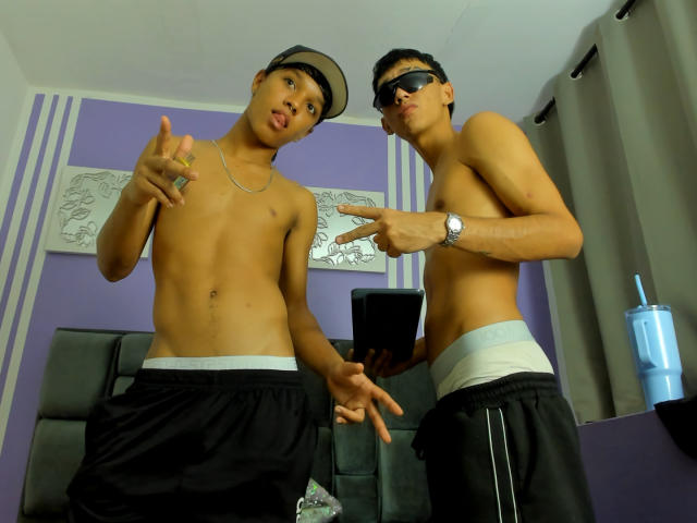 Two shirtless NickStebaldos with black pants and hats, making peace and rock signs, wearing sunglasses, standing in a room with purple walls and striped wallpaper.