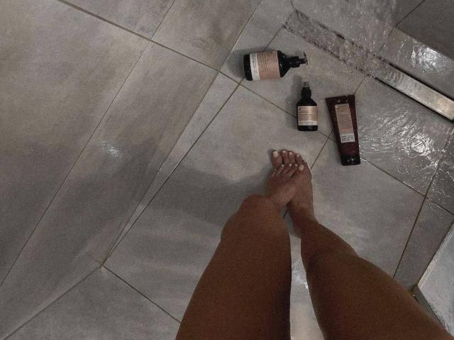 Los pies descalzos de MissiSofiya, con las uñas de los pies pintadas de blanco, están sobre el suelo de baldosas grises, al lado de dos botellas y un tubo marrón en el lado derecho..