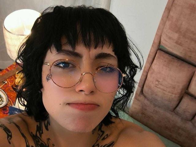 CleoZapata, avec ses cheveux noirs ondulés, ses yeux bleus et ses lunettes rondes, montrant des tatouages sur son cou et sa poitrine, nue, souriant légèrement, assise près d'une lampe..