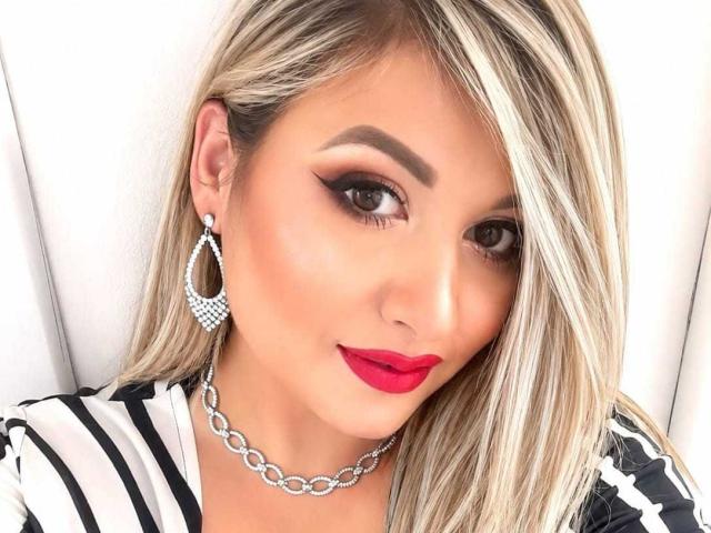 DalidaThierry with blonde hair, red lipstick, black eyeliner, diamond earrings, and chain necklace, showing off her sexy face and white shirt with black stripes.