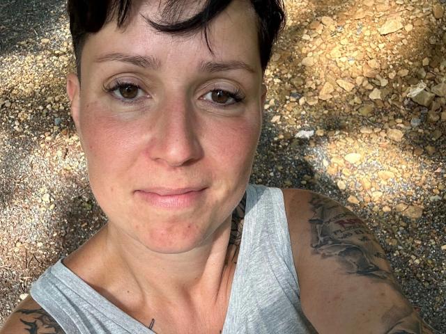 A TattoogirlDD-hot tem cabelo curto e escuro, olhos castanhos e pele clara com tatuagens nos ombros. Ela está vestindo uma regata cinza e está sorrindo levemente para a câmera..