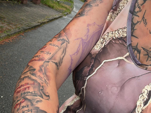 Il braccio di TattoogirlDD-hot è ricoperto di tatuaggi scuri, tra cui uno squalo e delle scritte, e indossa un abito viola, bagnato e trasparente, con pizzo, che mette in mostra il suo seno grande e umido. Sullo sfondo, si vede una strada..