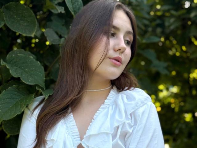 TessaLove-hot has long brown hair, white ruffled shirt, light skin, and a subtle, pouty expression with pink lips, standing against green leaves. She's wearing a thin white necklace.