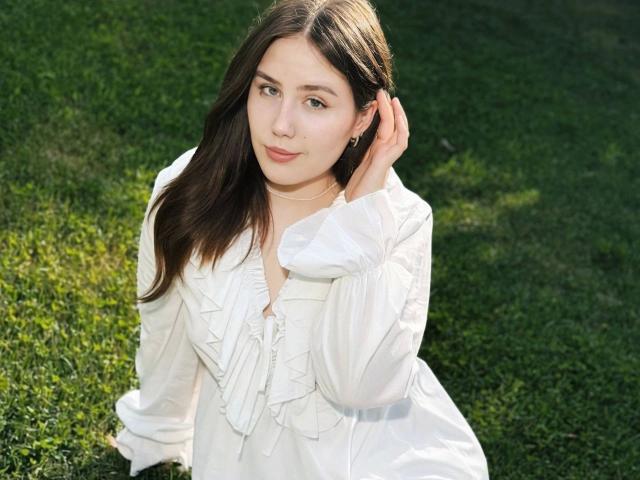 TessaLove-hot with long brown hair, wearing a white dress, sits on grass, touching her hair with a hand, showing a subtle smile, small tits, and a delicate necklace.
