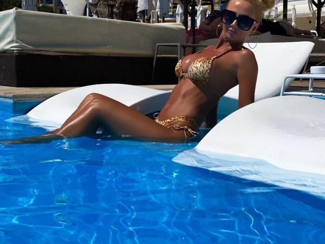 MoniqueBliss lounges in a pool, wearing a leopard-print bikini, her tan skin glistening, legs in blue water, white inflatable under her, sunglasses on, blonde hair up.
