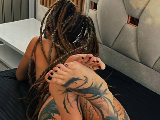 LucyDollyHot deitada em uma cama, de costas para a câmera, mostrando seus pés tatuados com esmalte preto, e longos cabelos castanhos trançados, em uma pose relaxada e sensual..