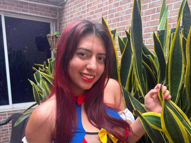 Brendaa Pierce, com cabelo ruivo, sorrindo, segurando folhas de planta-cobra, vestindo uma blusa azul com um laço vermelho e amarelo, mostrando seus seios, batom rosa, com um fundo de parede de tijolos..