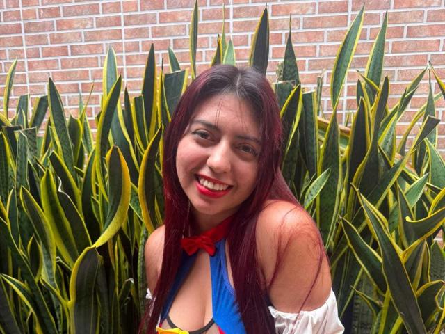 BrendaaPierce with long red hair, smiling, wearing a blue and red top with white sleeves, showing cleavage, surrounded by green and yellow snake plants.