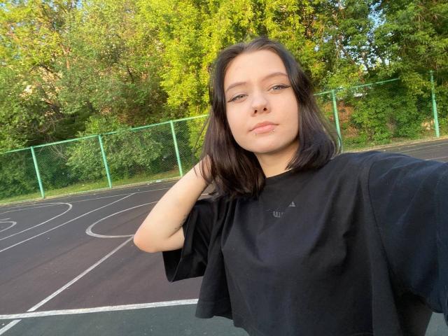 MikaMoony has pale skin, black shoulder-length hair, and is wearing a black t-shirt. She's taking a selfie on a tennis court with a green fence in the background.