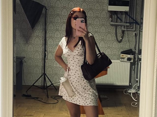 AlexXas, avec une poitrine de taille moyenne, portant une robe blanche avec des pois rouges, tenant une bouteille, un sac noir et un téléphone rose, debout devant un miroir, sur un sol en bois..