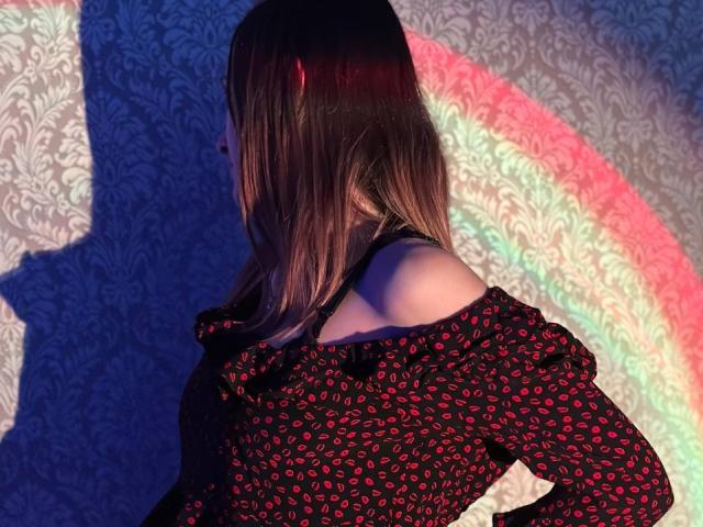 Seliina with red-patterned black dress, off-shoulder, shadow on wall, rainbow light, brown hair, back to camera, slight side view, standing, light blue patterned wall.
