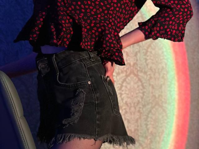 Seliina's red, black-patterned blouse with ruffled sleeves, and her frayed, black denim skirt with worn-out patches, standing with hands on hips, visible from the side.