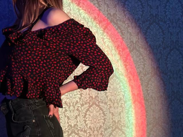 Seliina with red lip print black off-shoulder top, black jeans, hand on hip, standing against colorful rainbow light on patterned wall. Her ass is slightly visible.