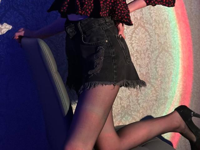 Seliina's black denim skirt with frayed edges, black sheer stockings, black heels, and red patterned top, leaning on a grey chair, showing her ass and legs.