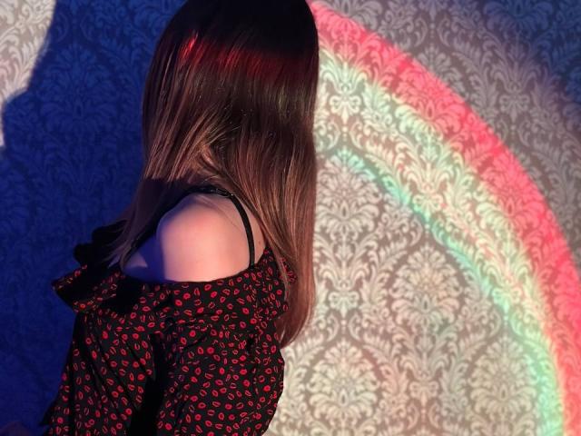 Seliina with straight brown hair, wearing a black off-shoulder dress with red lip prints, stands in a colorful rainbow shadow. Her back is to the camera.