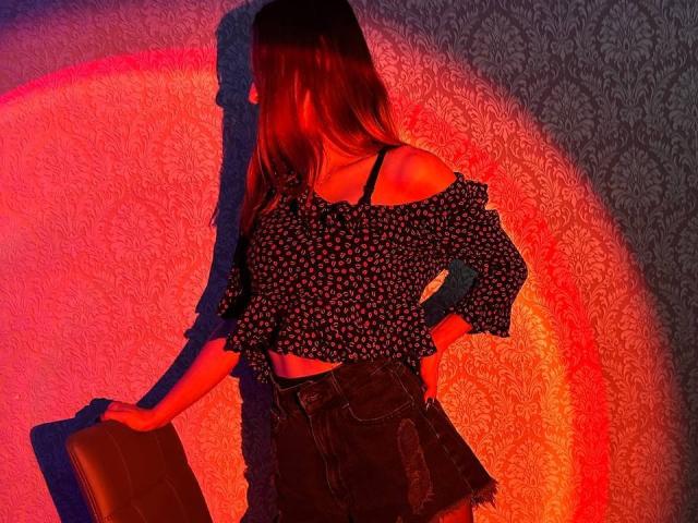 Seliina with long brown hair, black off-shoulder floral top, black bra strap visible, and black denim skirt, holding brown suitcase, shadowed by red and orange lights.