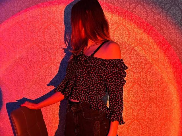 Seliina with long brown hair, black off-shoulder floral top, black straps, dark jeans, standing with hand on brown chair, shadowed by red-orange light.