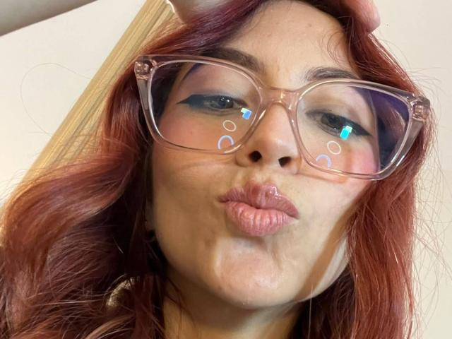 MiaaRouge with red hair, puffy pink lips, and transparent glasses, puckering up. Reflections of blue and white circles on her glasses. Her face is slightly wet.