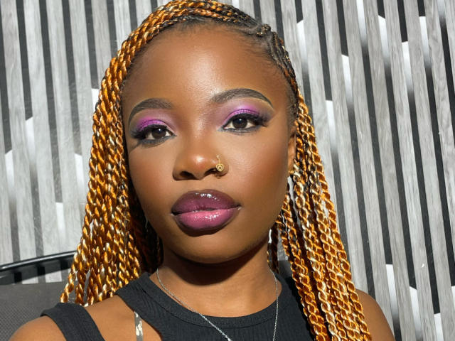 EbonyEmma tem a pele marrom escura, cabelo trançado laranja, maquiagem roxa nas pálpebras, batom rosa brilhante, um piercing no nariz, uma blusa preta e uma expressão séria. Suas nádegas não estão visíveis..