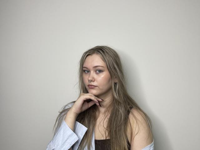 JonAzzara, avec de longs cheveux blonds et en désordre, portant une chemise blanche par-dessus un haut marron, le menton posé sur la main, avec une expression sérieuse, une peau pâle, des yeux bleus, un petit anneau de nez, et une expression neutre..