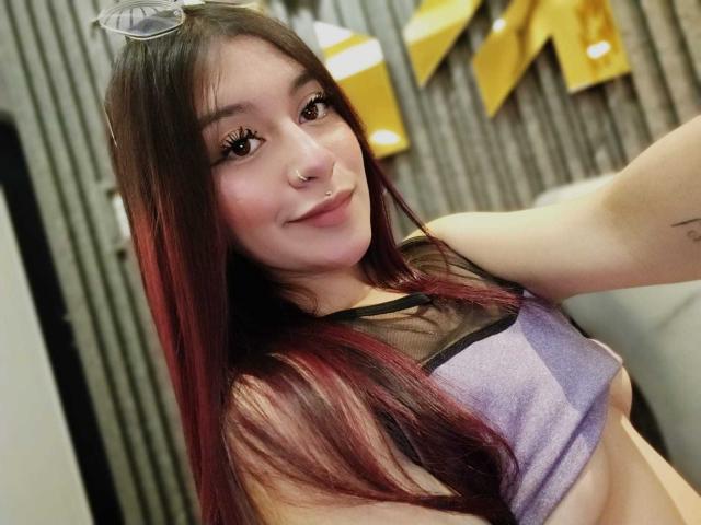 AngelsPhoenix with long red-brown hair, nose ring, and eyelashes, wearing a purple and black mesh top, smiling, showing her tits, and wearing glasses in a selfie.
