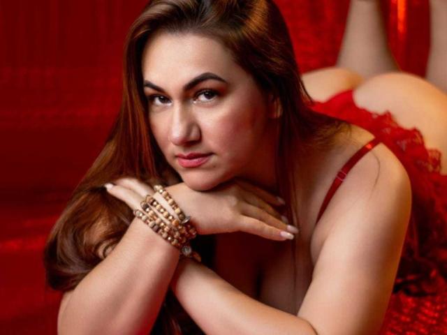 AlmaMature with red lace lingerie, lying on her stomach, showing her ass, with brown hair, red eyes, and a bracelet, looking at the camera with a seductive expression.