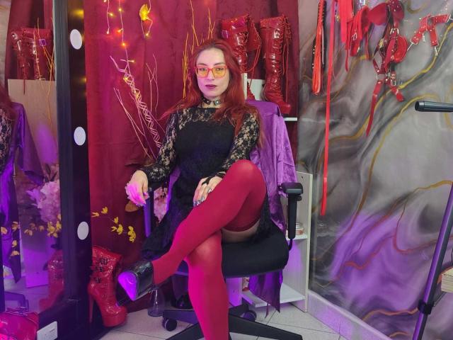 QueenScarletSecret, com cabelo ruivo, óculos, vestido de renda preta, meias vermelhas até a coxa e saltos pretos, sentada em uma cadeira com iluminação roxa, cercada por botas vermelhas e equipamentos BDSM..