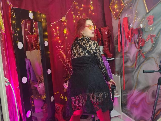 QueenScarletSecret com óculos vermelhos, vestido de renda preta e botas vermelhas, em frente a um espelho com luzes, cercada por luzes de cordão vermelhas e amarelas..