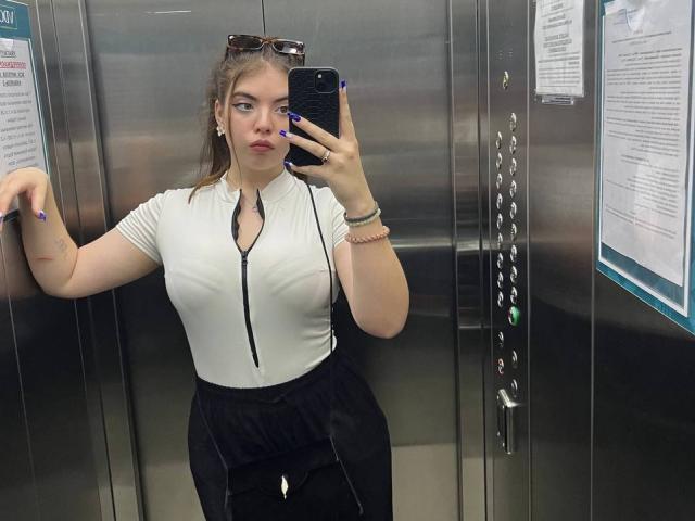 MagdaLia macht ein Selfie vor einem Spiegel in einem Aufzug, trägt ein weißes, vorne zuziehendes Hemd, das ihre Brüste zeigt, schwarze Hosen und blauen Nagellack, und hat eine Sonnenbrille auf dem Kopf..