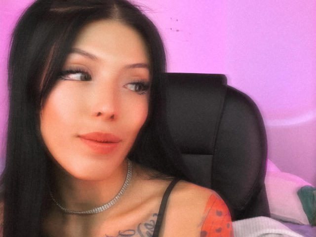 AizenMyoo tiene el cabello largo y negro, piel clara y un tatuaje en el hombro derecho. Lleva un top de tirantes negros y un collar plateado, y está mirando hacia un lado..