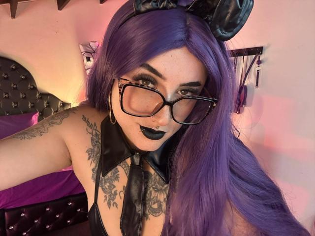 LilithDark69 with purple hair, black glasses, black lipstick, black leather bunny ears, and tattoos, wearing a black leather halter, showing off her tits and a nose ring.