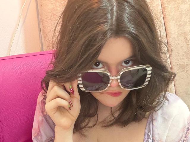 AlexaMissFortune with wavy brown hair, wearing striped sunglasses, pink nails with red and purple tips, and a pinkish-purple top, looking at the camera with red lips.