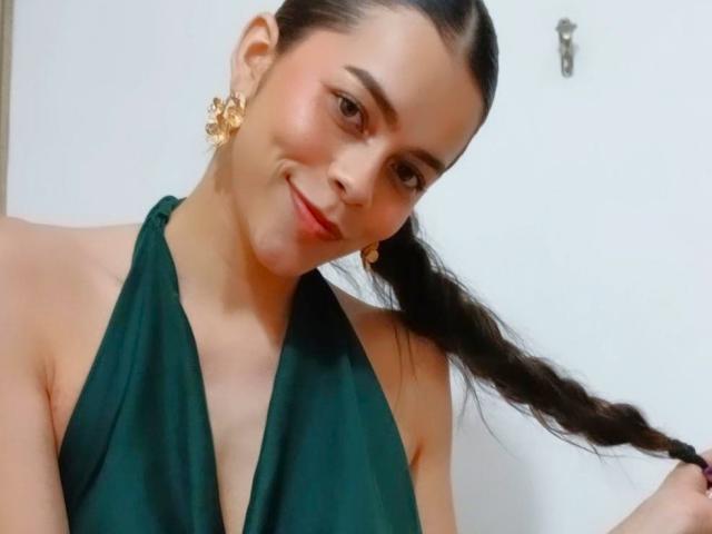Alana Blake tem cabelo escuro trançado, um vestido verde de alças, brincos de flor dourados e um sorriso discreto. Ela está segurando a trança com uma expressão adorável e relaxada..