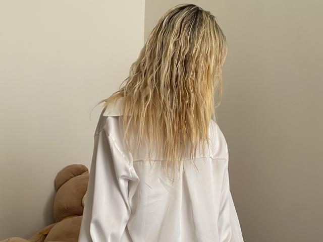 MysteryLaddy with wavy blonde hair, wearing a white oversized shirt, back to camera, standing next to a beige stuffed teddy bear.