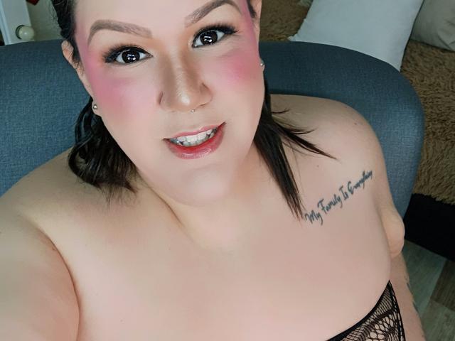 GiiannaMorrys with pink blush, black eyeliner, and nose ring, showing teeth, has tattoos on right shoulder, and is wearing black lace, looking up at camera.