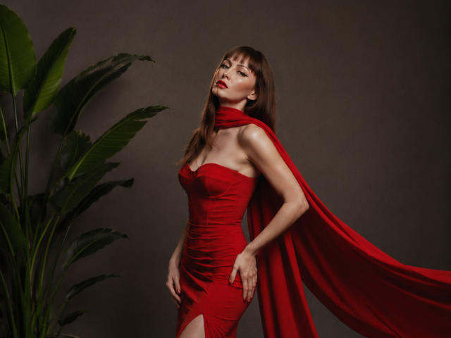 MiraMixi, avec une robe rouge, une cape et de longs cheveux bruns, debout, regardant vers le haut, rouge à lèvres rouge, plante verte sur la gauche, mains sur les hanches, sexy, mince, petites seins, fond sombre..