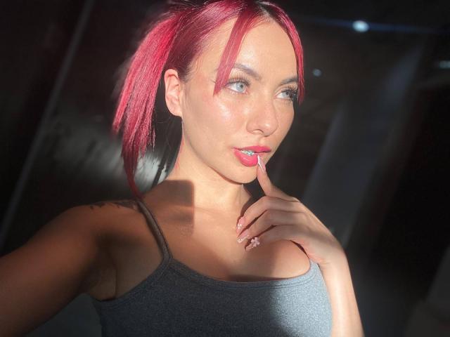 Valery Harllow mit roten Zöpfen, einem grauen Tanktop, glatter Haut, einem Finger, der an den rosafarbenen Lippen ruht, einem Tattoo auf der Schulter, Sonnenlicht, das das Gesicht hervorhebt, blauen Augen und rosafarbenem Nagellack..