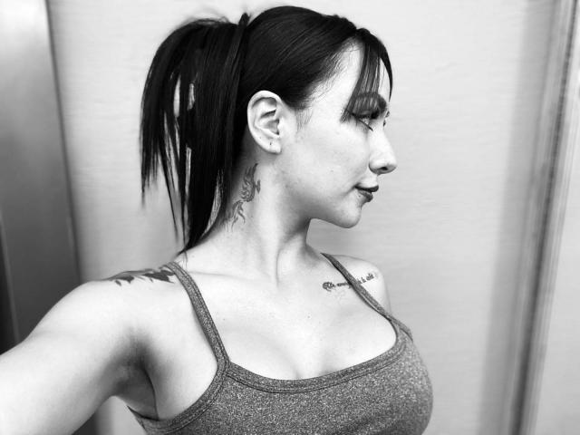 ValeryHarllow with dark ponytail, gray tank top, tattoos on neck and shoulder, medium boobs, side profile, black and white, ear piercings, looking right, slight smile.