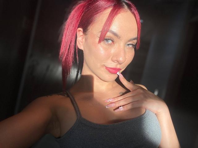 ValeryHarllow with pink hair in a ponytail, wearing a black tank top, sunlight highlighting her face, pink lipstick, and finger touching her chin, showing off her shiny nails.