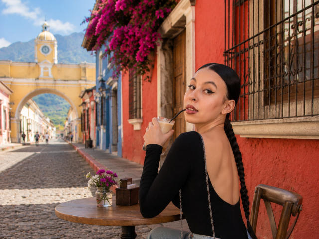 NenaBlack with long black braid, black long-sleeve top showing her ass, sipping from a straw, sitting at a wooden table with flowers, in a colorful cobblestone street.