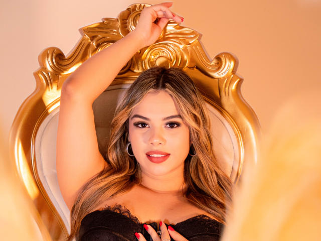 ZoeVibe with her arm raised, sitting on a gold, ornate chair, wearing black lace, showing her tits, red nails, and smiling with hoop earrings. Blonde hair, light skin.